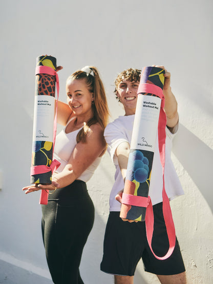 Two people holding rolled Wild Mingo Pink Lady Yoga Mats with a white stucco wall behind